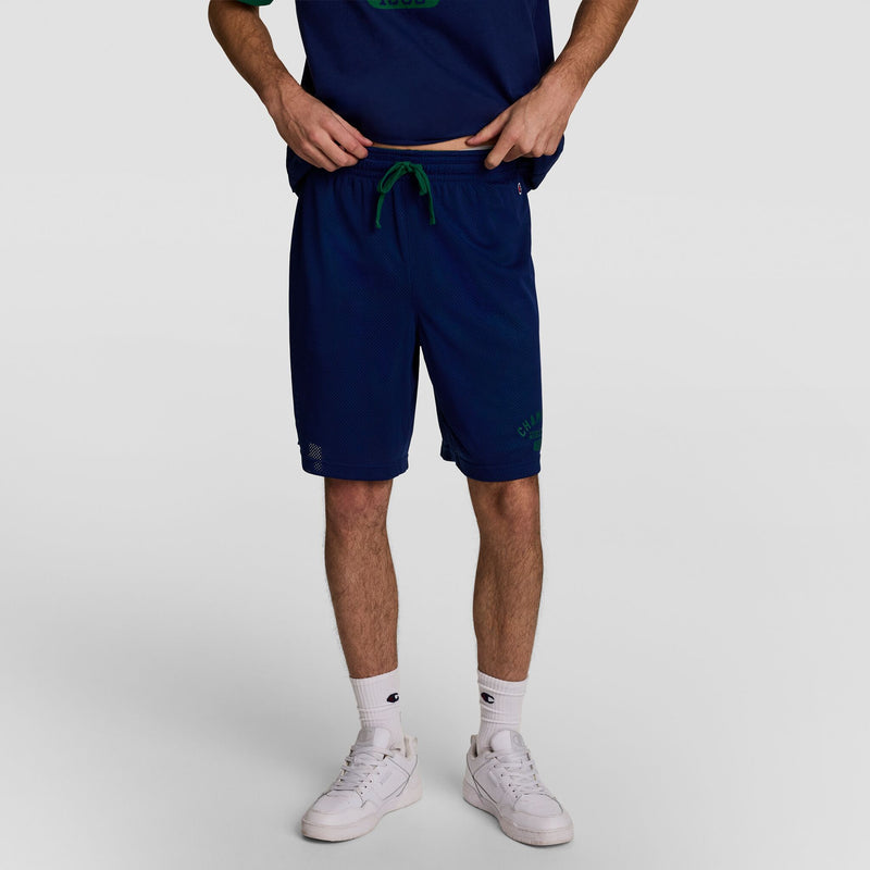 A person wears Champion’s Big & Tall 8" Fleece Cargo Short with C Logo in navy, paired with a matching navy shirt, white socks, and white sneakers. They stand against a plain white background; their head is not visible.