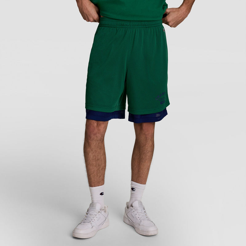 A person stands against a plain light background, wearing Champion 8" Fleece Cargo Short, C Logo - Big & Tall in green layered over blue shorts, with white socks and sneakers. The upper half of their body is cropped out.