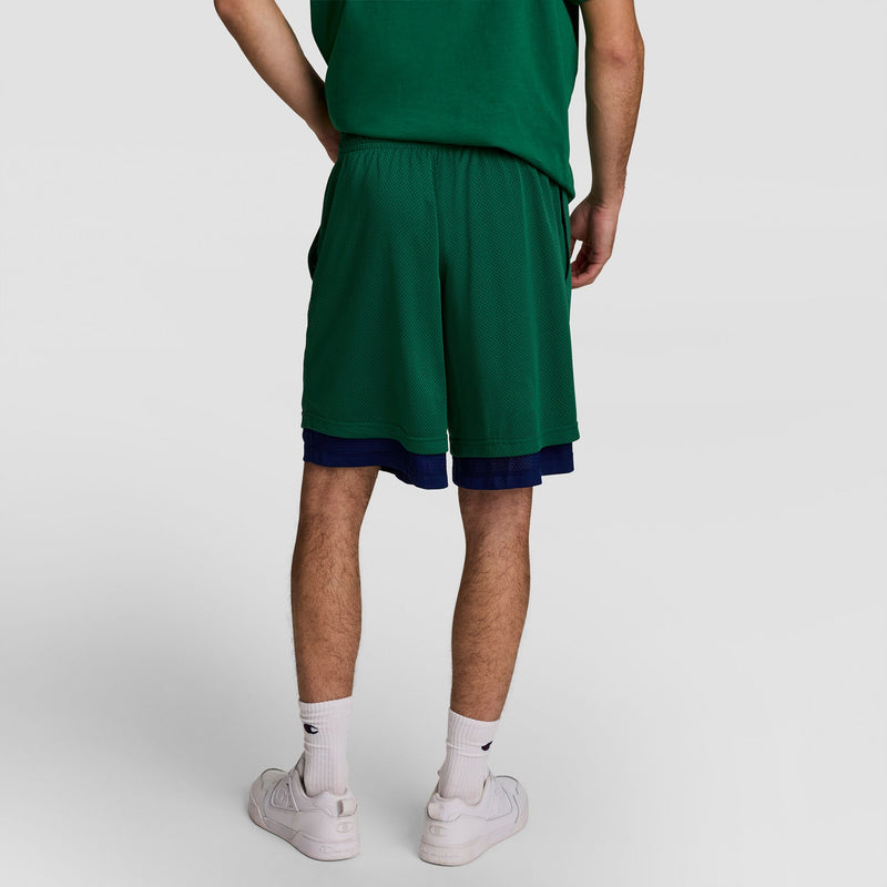 A person faces away wearing a green T-shirt, Champion 8" Fleece Cargo Short, C Logo - Big & Tall over blue shorts, white crew socks, and white sneakers. The background is plain and neutral.