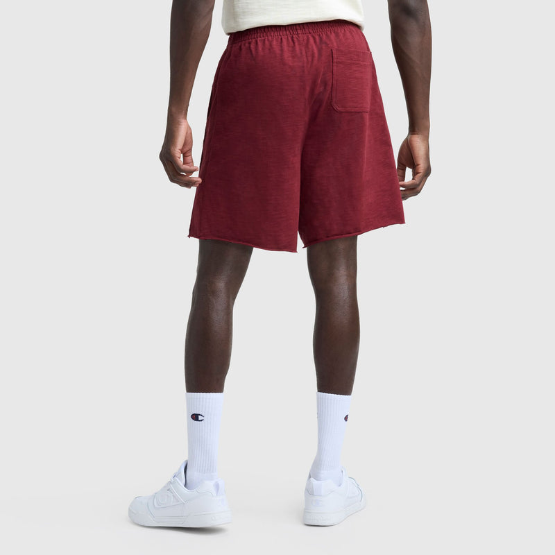 A person stands facing away in a cream shirt, Champion 9" Modern Retro Washed Shorts with C Logo in maroon featuring a back pocket, white Champion crew socks, and white sneakers against a plain light background.