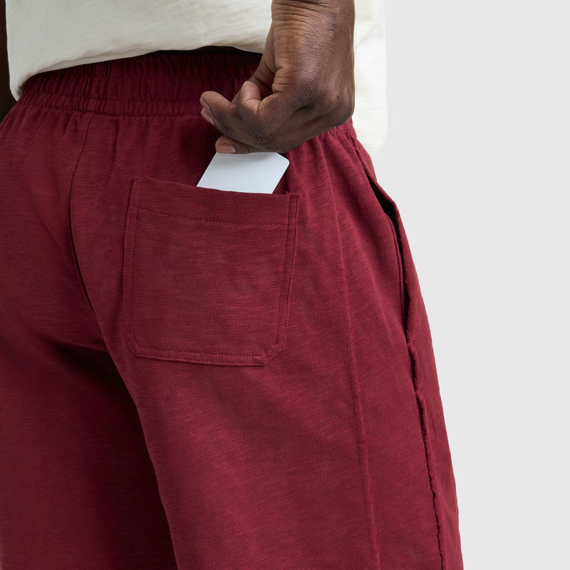 A person wearing Champion 9" Modern Retro Washed Shorts with the C Logo slips a white card into the back pocket, hand partially in, against a plain light gray background.