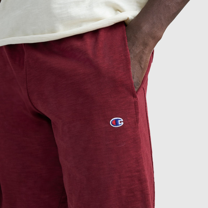 Someone wearing the Champion 9" Modern Retro Washed Shorts in maroon, with the C logo visible on the left thigh and a hand in the pocket; a cream shirt is partially visible.
