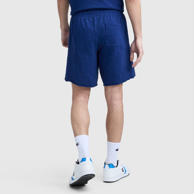 A person faces away, wearing a blue T-shirt, Champion 9" Modern Retro Washed Shorts with C Logo and back pocket, white Champion socks, and white sneakers with blue details against a plain light background.