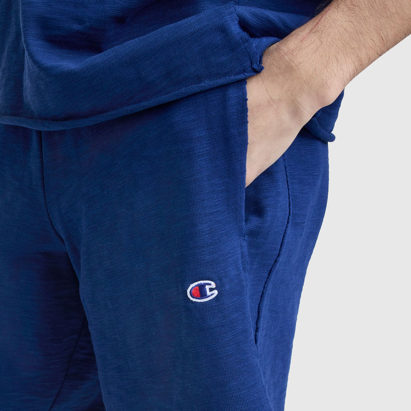 A person wears Champion's 9" Modern Retro Washed Shorts with a matching blue top, placing their right hand in a pocket. The shorts feature an embroidered C logo near the pocket for a sporty accent.