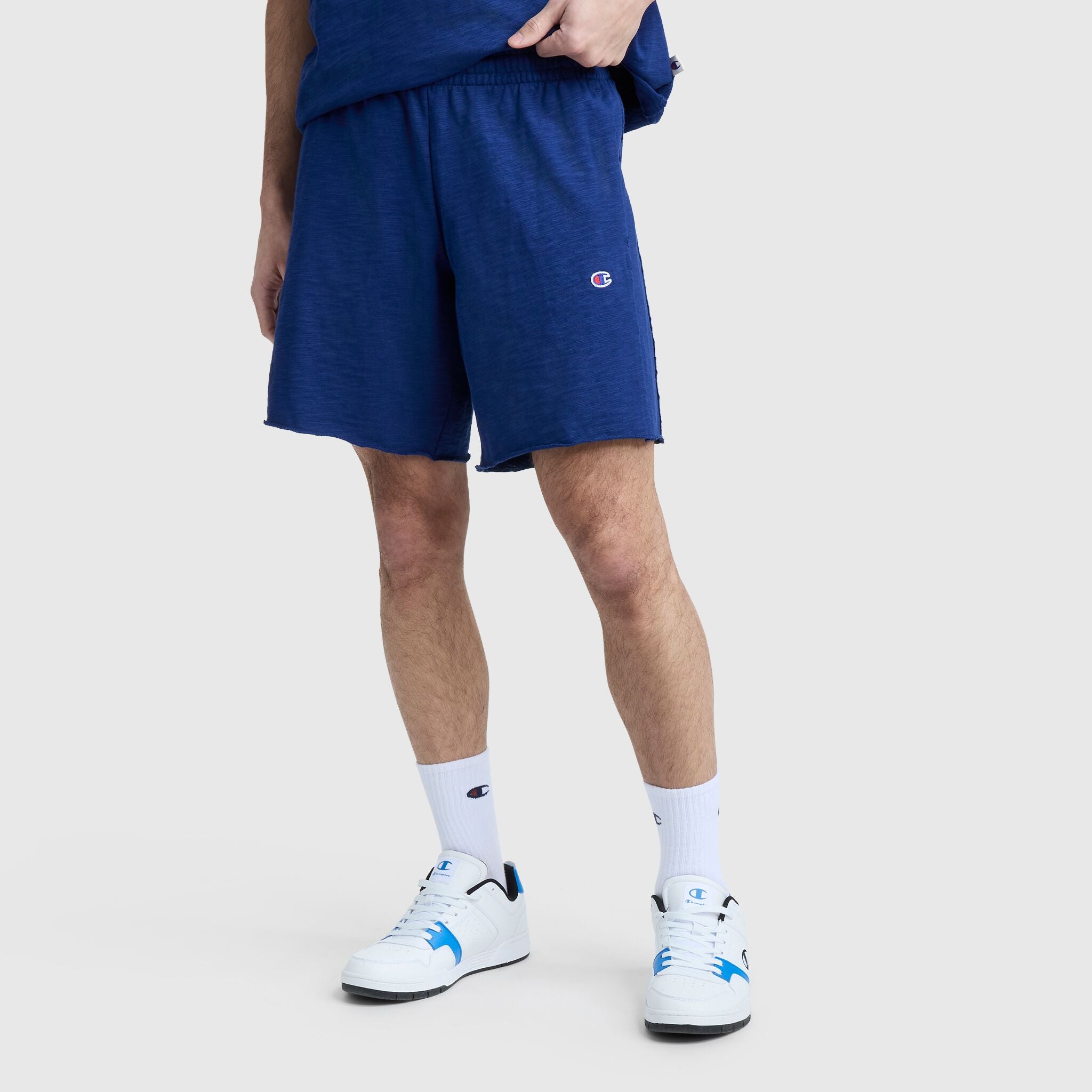Wearing Champion’s 9" Modern Retro Washed Shorts with C Logo in blue, this person pairs them with a matching blue tee, white Champion socks, and white sneakers featuring blue accents. Their face and upper body are not shown.