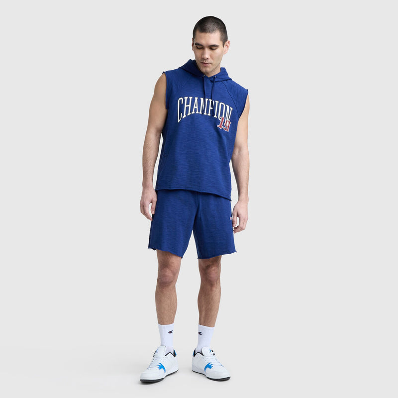 A man in a sleeveless blue hoodie with CHAMPION on the front, Champion 9" Modern Retro Washed Shorts with C Logo, white crew socks, and white athletic shoes stands against a plain light gray background.