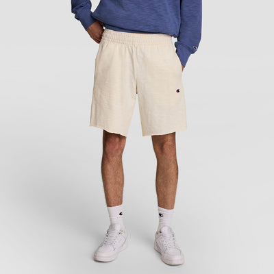 A person wears Champion 9" Modern Retro Washed Shorts with C Logo in cream, paired with a blue sweatshirt, white crew socks, and white sneakers, standing against a plain background with hands in their shorts pockets.