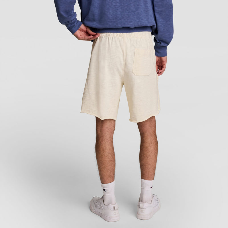 A person stands facing away, wearing a blue long-sleeve shirt and Champion 9" Modern Retro Washed Shorts, C Logo, in cream cotton with a back pocket, paired with white crew socks and white sneakers against a plain light background.