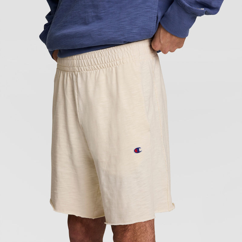 A person wears cream 9" Modern Retro Washed Shorts with an elastic waistband and Champion’s C logo on the left leg, paired with a blue sweatshirt, shown from waist to mid-thigh.