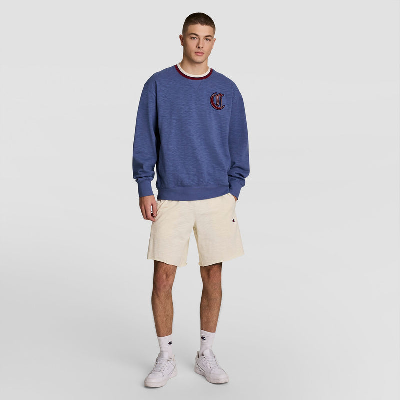 A young man with short hair and a neutral expression wears Champion 9" Modern Retro Washed Shorts with C Logo in cream, a blue sweatshirt with a small emblem, white socks, and white sneakers against a plain background.