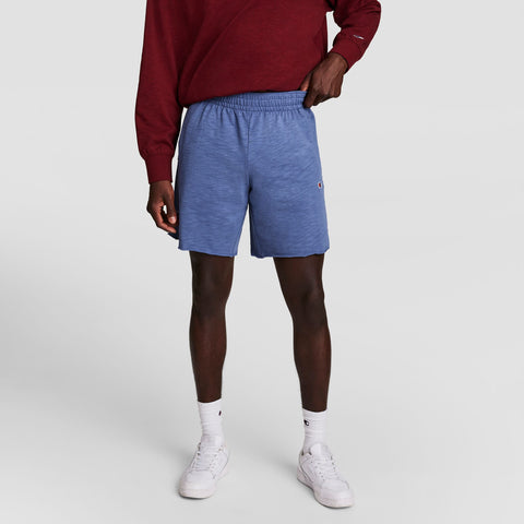 A person wearing a maroon long-sleeve shirt, Champion’s 9" Modern Retro Washed Shorts with C Logo in blue, white crew socks, and white sneakers stands with one hand in their pocket against a plain background.