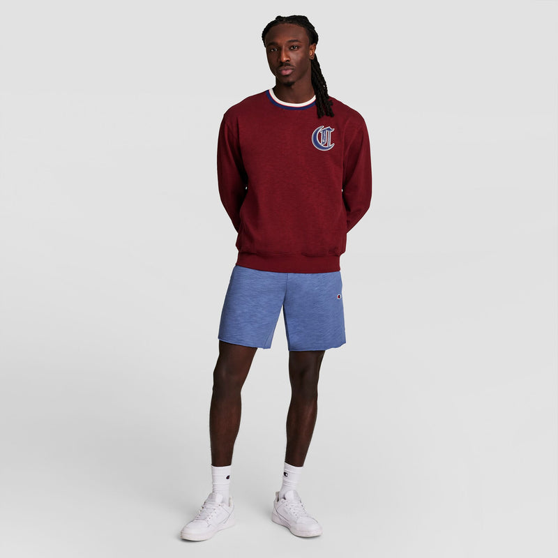 A man stands against a plain light background wearing a maroon sweatshirt, Champion 9" Modern Retro Washed Shorts with C Logo in blue, white crew socks, and white sneakers. His hands are behind his back.
