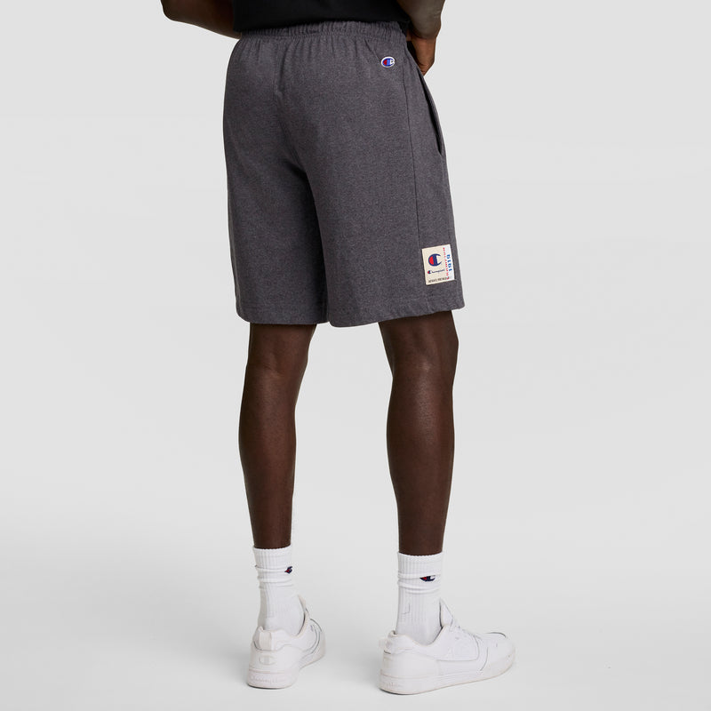 A person stands facing away, wearing Champion 7" Jersey Short with C Logo in dark gray, paired with white crew socks featuring a small logo and white sneakers against a plain light background.