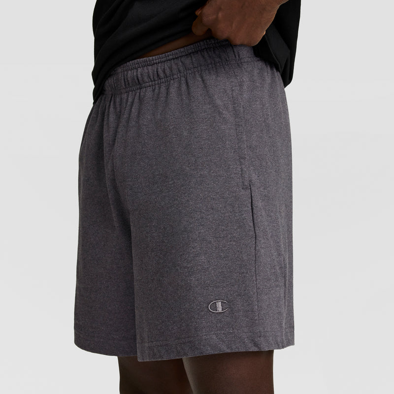A person wearing Champion 7" Jersey Short, C Logo in dark gray, featuring an elastic waistband and small logo near the hem, stands against a plain light background.