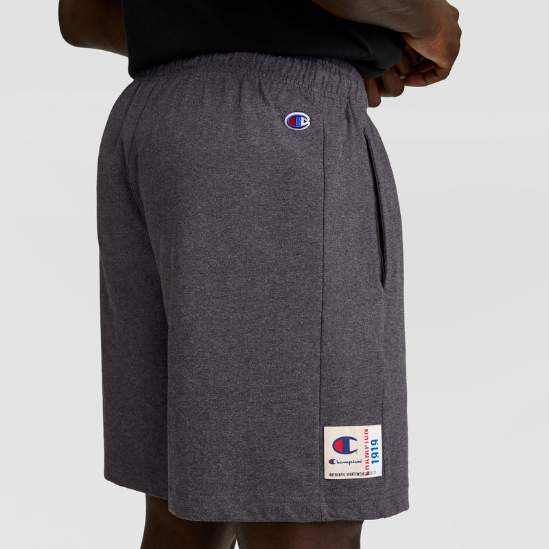A person wears dark gray Champion 7" Jersey Shorts, featuring the C logo on the left thigh and a small patch near the hem, with hands on hips against a plain light gray background.