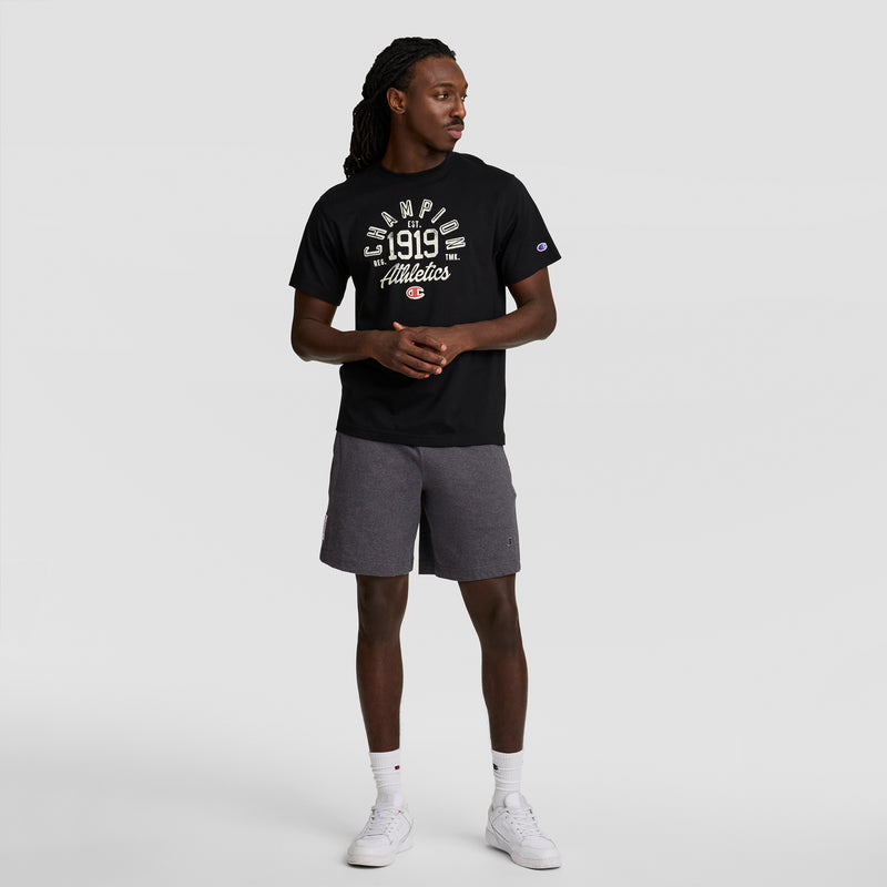 A man stands against a plain background wearing a black Champion 1919 Athletics t-shirt and Champion 7" Jersey Short with C Logo in gray, paired with white sneakers and socks. He looks left with his hands clasped in front of him.