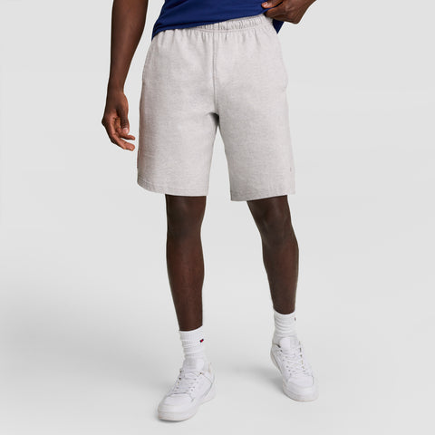 A person is shown from the waist down wearing Champion 7" Jersey Shorts with the C logo, paired with white socks and sneakers, standing against a plain light background with one hand in their pocket.