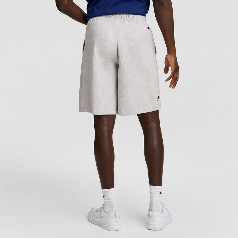 A person stands facing away, wearing Champion 7" Jersey Short with C Logo in light gray, paired with a blue top, white socks, and white sneakers against a plain light background.
