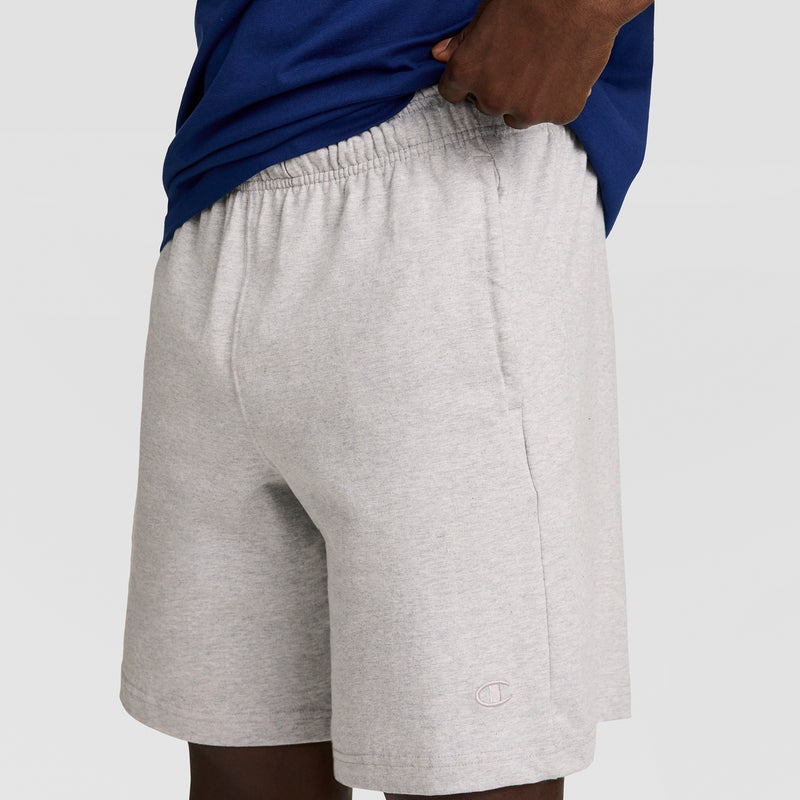 A person is wearing Champion 7" Jersey Shorts with an elastic waistband and a small C logo near the hem; their hand is in a pocket, with only the lower torso and upper legs shown.