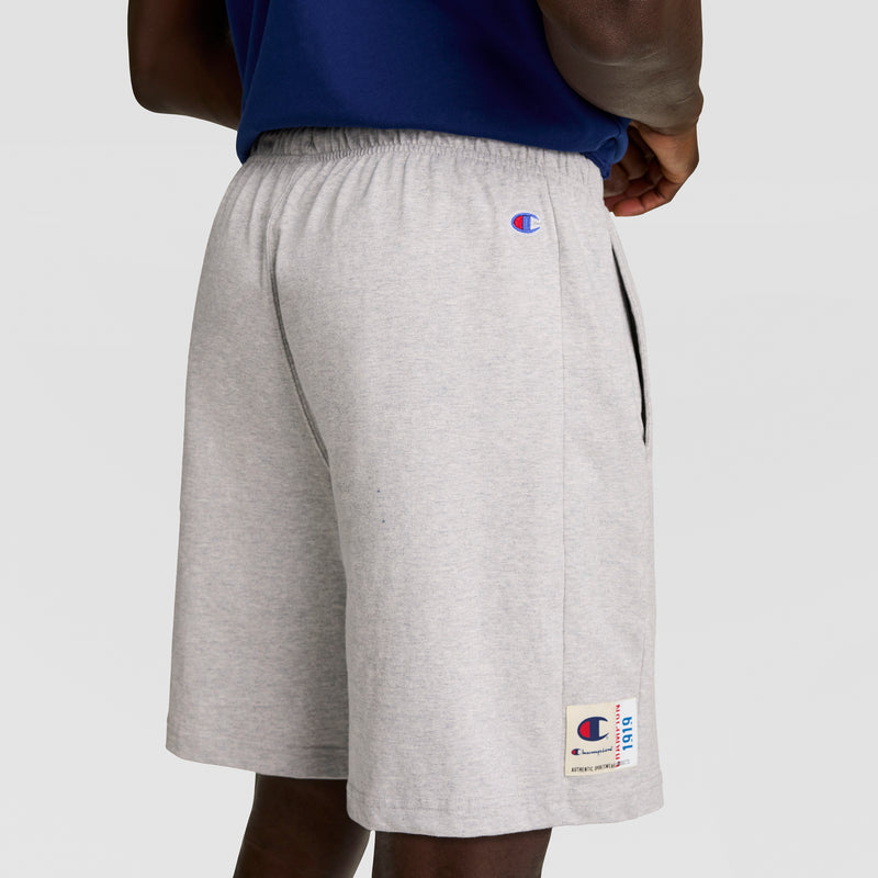 A person wears light gray Champion 7" Jersey Shorts with an elastic waistband, side pockets, and visible C logo, paired with a navy blue shirt. The shorts are cotton jersey. The background is plain and light.