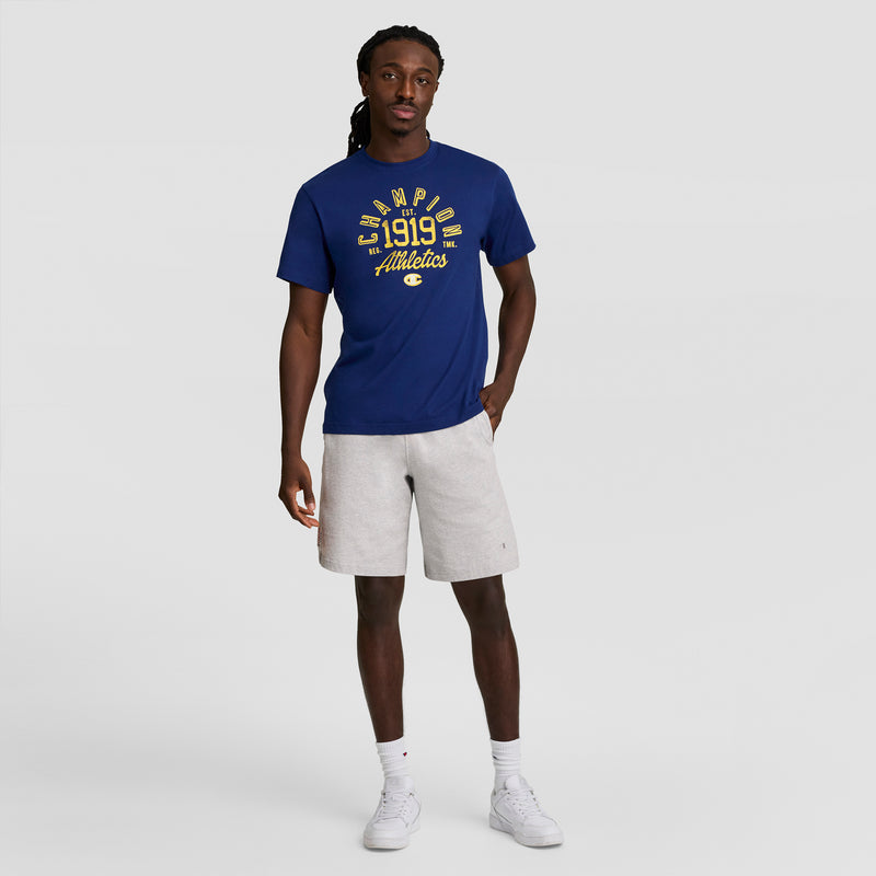 A man faces forward in a blue Champion 1919 Athletics T-shirt, light gray Champion 7" Jersey Shorts with C Logo, and white sneakers against a plain light gray background.