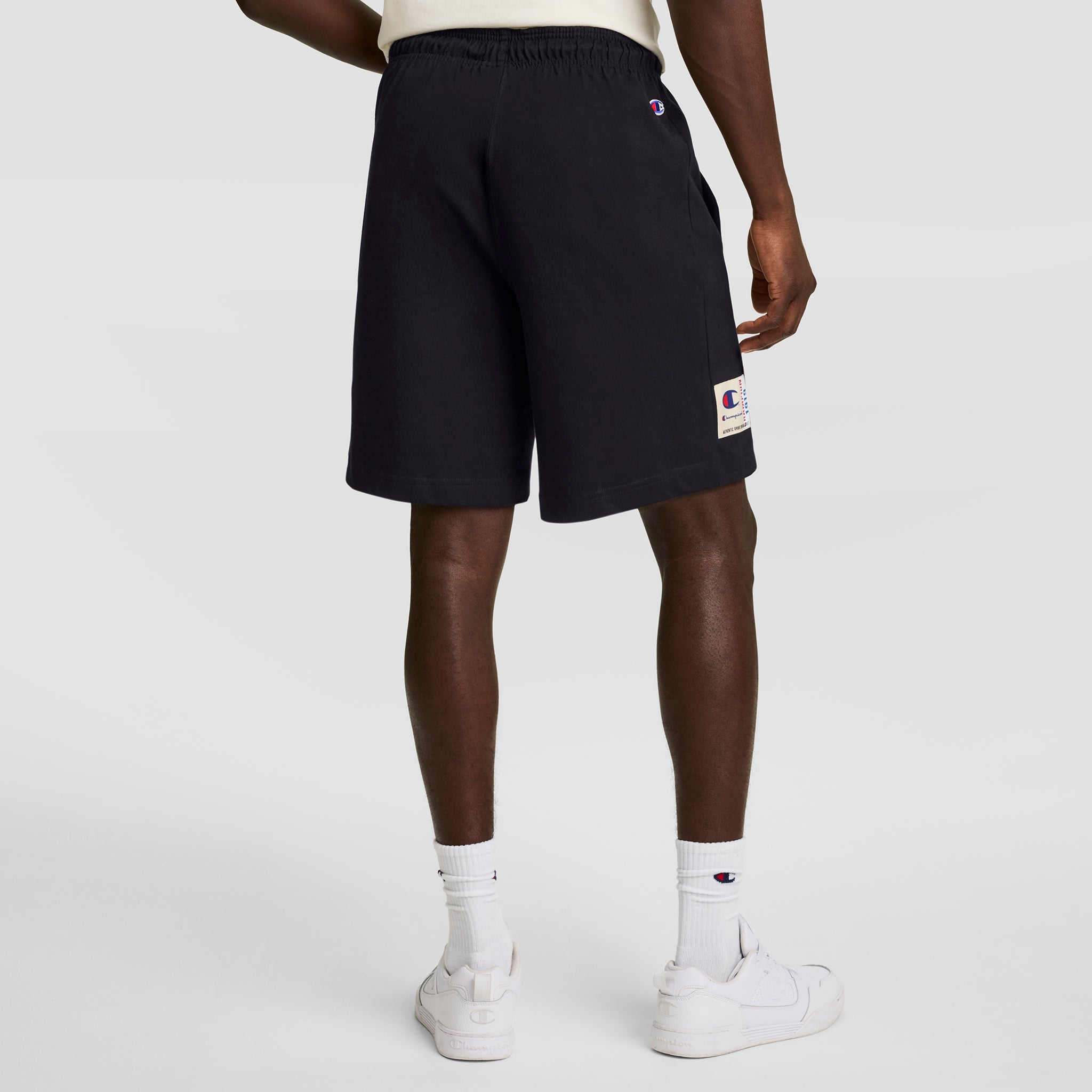 A person faces away, wearing Champion 7" Jersey Short with a C logo patch, paired with white socks and sneakers. The background is plain and light-colored.