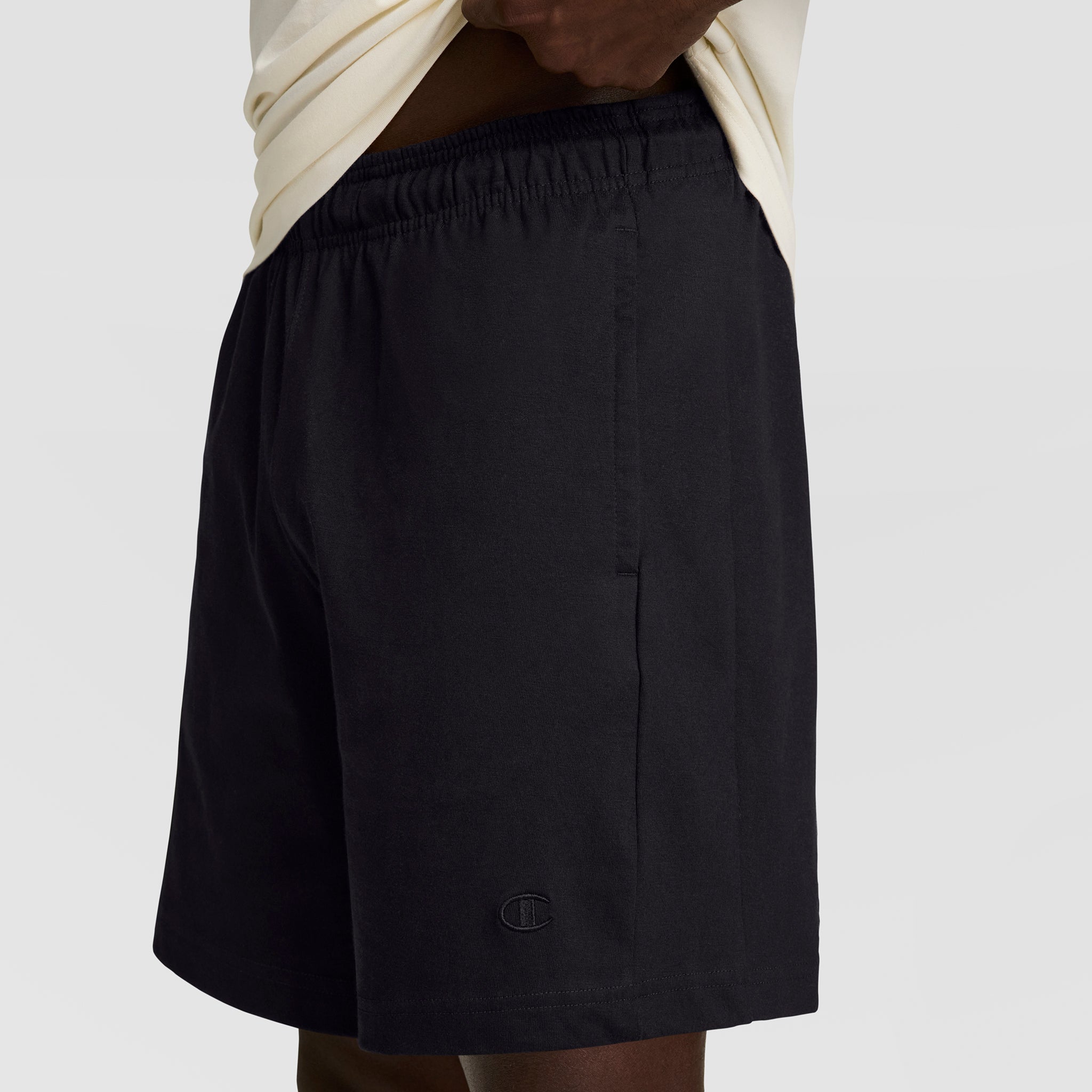 A person in a light shirt lifts it to reveal Champion's 7" Jersey Short, C Logo—black men’s shorts made from soft cotton jersey with an elastic waistband and a small embroidered logo by the hem for classic Champion style.