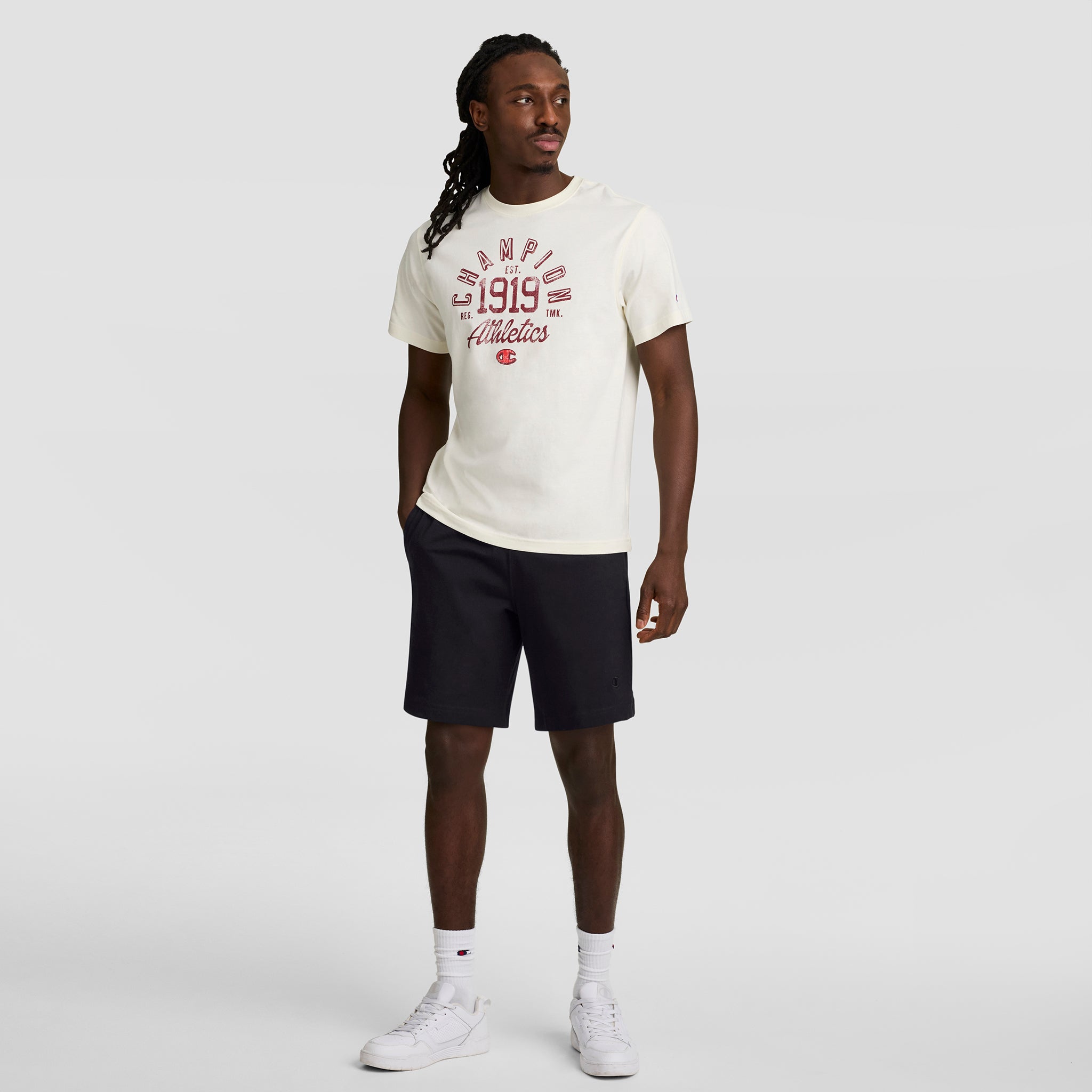 A man with long hair stands against a plain background in a white cotton Champion Athletics 1919 T-shirt, Champion 7" Jersey Short with C Logo (black), white socks, and white sneakers, one hand in his pocket.