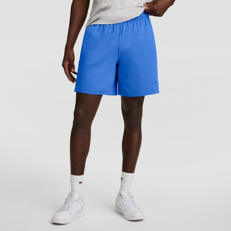 A person in a light gray t-shirt, white crew socks, white sneakers, and bright blue Champion 7" Jersey Shorts with C Logo stands against a plain light gray background.