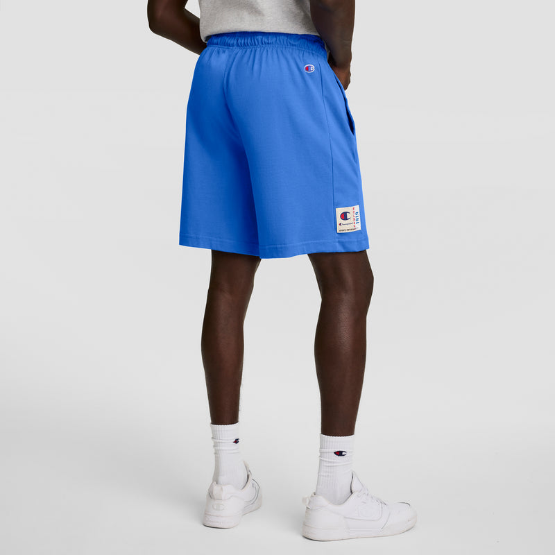 A person faces away, wearing a gray shirt, white sneakers and socks, and bright blue Champion 7" Jersey Shorts with the C logo; the Champion branding is visible on the shorts and socks against a plain background.