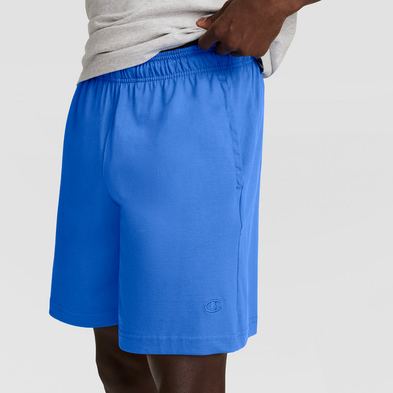 A person is wearing a light gray T-shirt and Champion's 7" Jersey Short, C Logo in bright blue with an elastic waistband, standing against a plain light background. Only their torso and upper legs are visible.