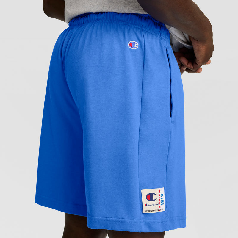 A person wearing Champion 7" Jersey Short, C Logo in bright blue, featuring logo patches on the side and near the pocket, stands with hands clasped behind their back.