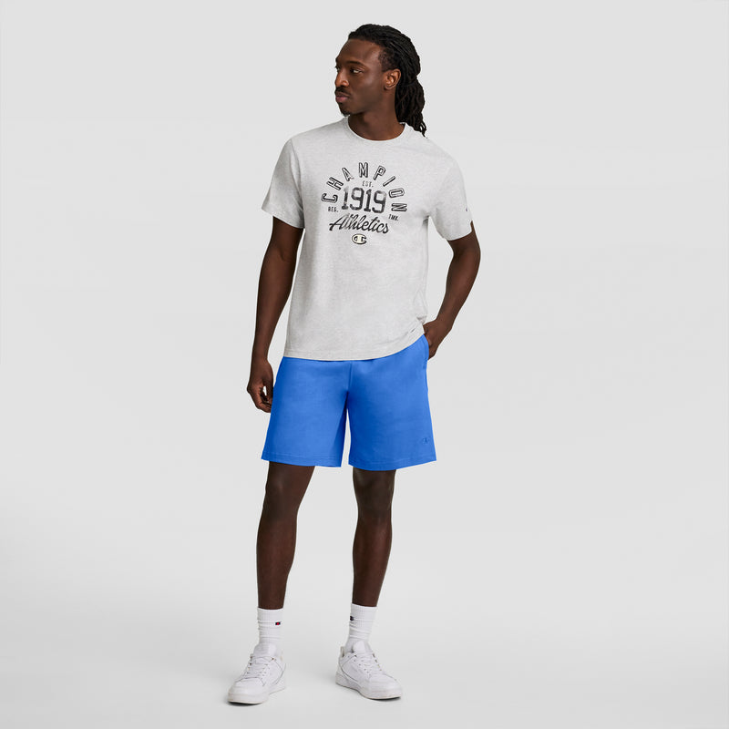A man with long hair stands against a plain background, wearing a light gray graphic t-shirt, white socks, white sneakers, and bright blue Champion 7" Jersey Short with C Logo. He looks left with one hand in his pocket.
