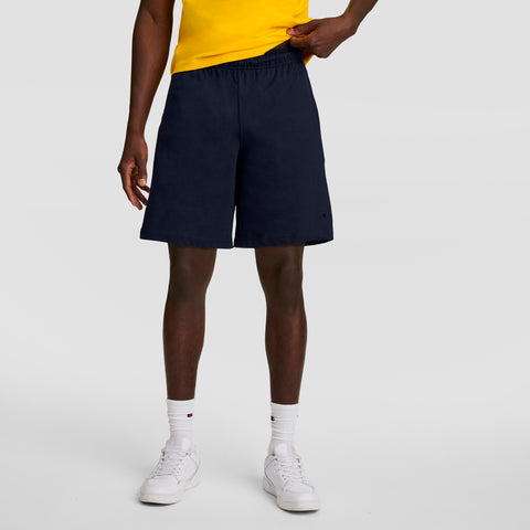 A person, visible from the shoulders down, stands against a plain light gray background wearing a yellow shirt, white socks, white sneakers, and navy blue Champion 7" Jersey Short with C Logo.