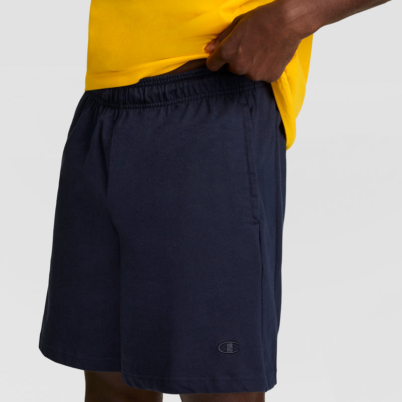 A person in a yellow shirt and dark blue Champion 7" Jersey Short, featuring an elastic waistband and C Logo on the left leg, stands against a plain light background.
