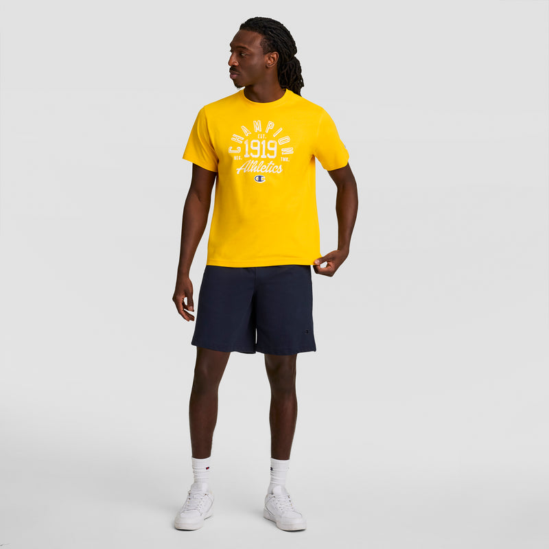 A man in a yellow Champions 1919 Athletics t-shirt, Champion 7" Jersey Short with C Logo in navy blue, white socks, and white sneakers stands against a plain light gray background, looking to his left.