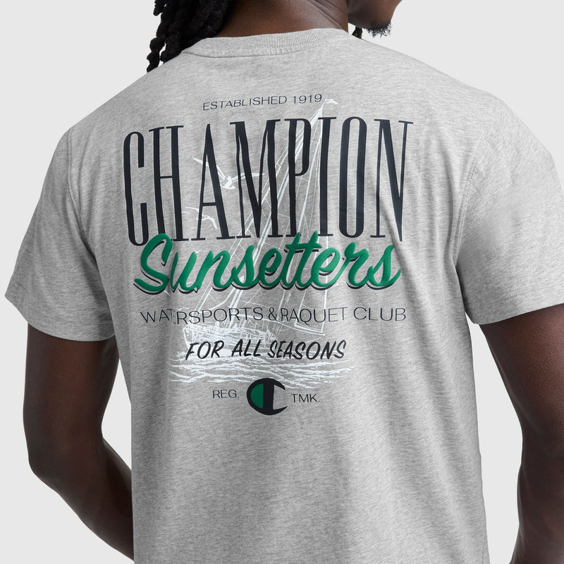 A person wearing a Champion Sunsetters Graphic T-Shirt, C Logo, in light gray with navy and green text on the back: CHAMPION Sunsetters WATSPORTS & RAQUET CLUB FOR ALL SEASONS ESTABLISHED 1919 REG. TMK.