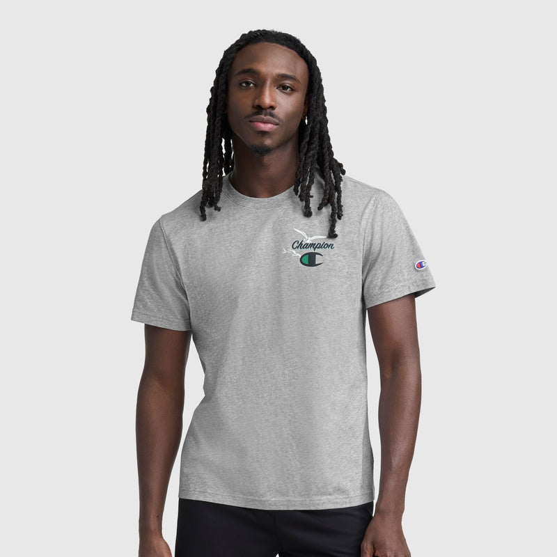 A man with long braided hair wears the Champion Sunsetters Graphic T-Shirt, C Logo—a light gray men's cotton tee with a small left chest logo and sleeve patch—standing against a plain light gray background.
