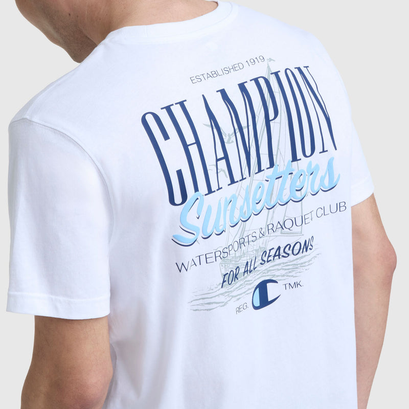 A person wearing the Champion Sunsetters Graphic T-Shirt, featuring blue and black text on the back that reads "Sunsetters Watersports & Raquet Club For All Seasons" along with smaller text and a stylized C logo.