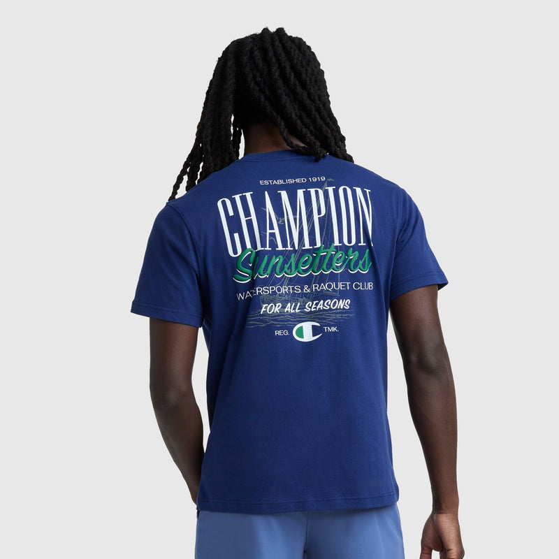 A person with long braids wears the Champion Sunsetters Graphic T-Shirt, C Logo—a men's cotton tee featuring "Sunsetters Watersports & Racquet Club For All Seasons" printed on the back in white, green, and blue.