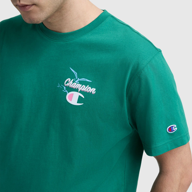 A person is shown from the shoulders down wearing a green Champion Sunsetters Graphic T-Shirt, C Logo, featuring a bird design and Champion branding on the chest and sleeve, against a plain background.