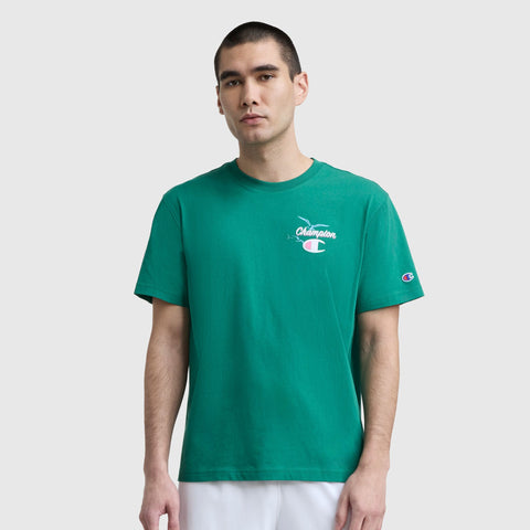 A young man with short dark hair wears a green Champion Sunsetters Graphic T-Shirt, C Logo, and white pants against a plain background. The tee features a small white Champion logo on the chest and sleeve.