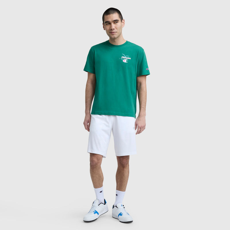 A man stands against a plain background wearing a Champion Sunsetters Graphic T-Shirt, C Logo in green, paired with white shorts, white crew socks, and white sneakers with blue accents.