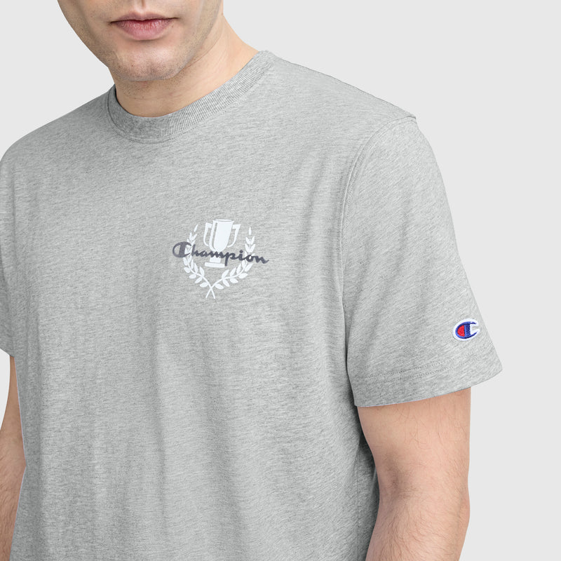 A person wears a light gray Champion Club Graphic T-Shirt, C Logo, with the Champion emblem on the chest and “C” patch on the left sleeve; only their lower face and upper torso are shown.