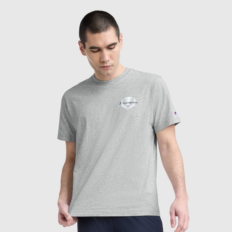 A young man wears a light gray Champion Club Graphic T-Shirt, C Logo by Champion, made from soft cotton jersey with a small white logo on the left chest. He stands against a light background, looking down with one hand by his hip.