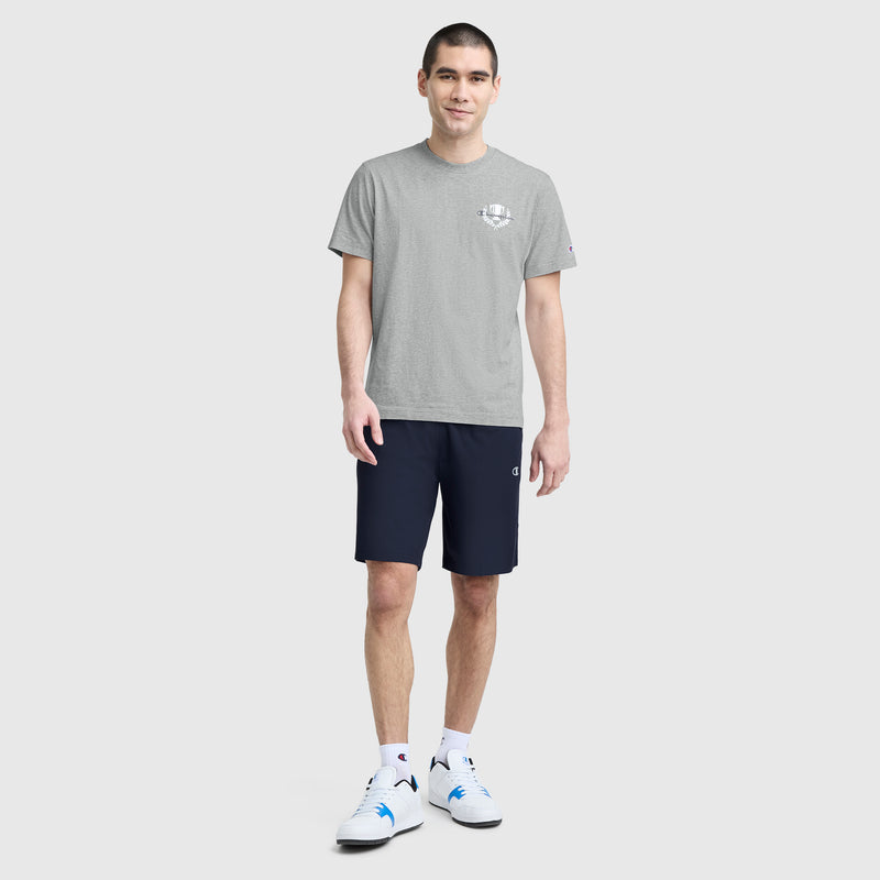 A man with short dark hair smiles against a plain background, wearing a Champion Club Graphic T-Shirt with C Logo by Champion, paired with navy shorts, white socks, and white athletic shoes.