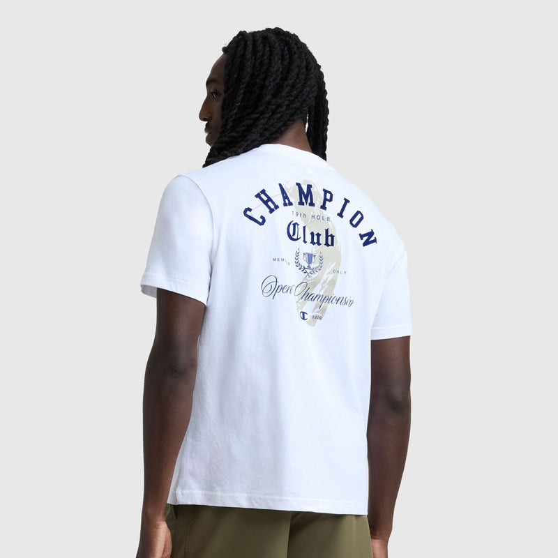 A person with long braids, facing away from the camera, wears a Champion Club Graphic T-Shirt, C Logo by Champion. The 100% cotton tee has blue and beige golf-themed graphics on the back and is styled with green shorts.