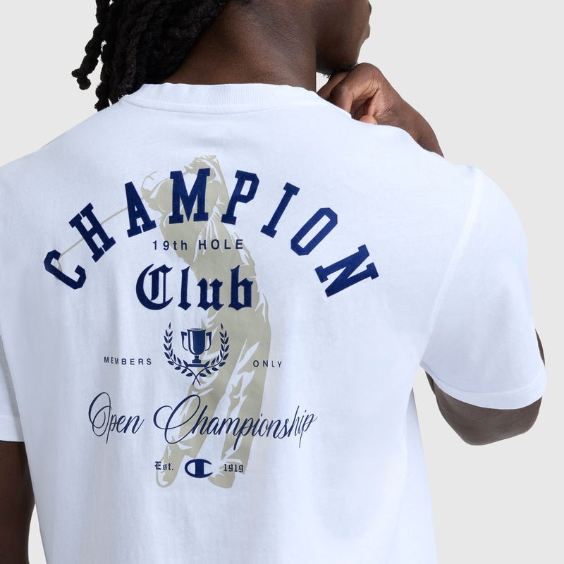 A person wears the Champion Club Graphic T-Shirt by Champion, made from 100% cotton, featuring blue "CHAMPION Club 19th Hole Open Championship" text, a trophy graphic, and a laurel wreath design on the back.