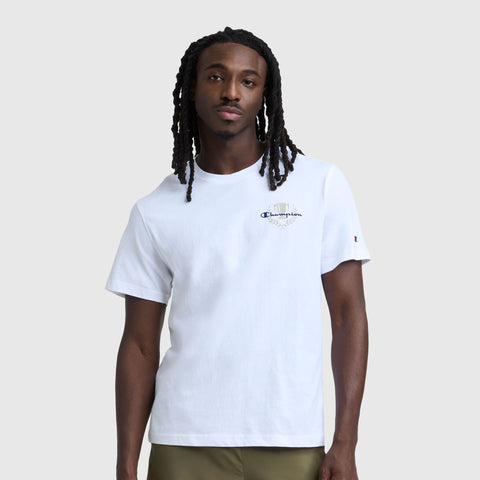 A man with long, braided hair wears a Champion Club Graphic T-Shirt, C Logo by Champion. The 100% cotton tee features a small embroidered logo on the left chest. He stands against a light gray background, facing the camera with a neutral expression.