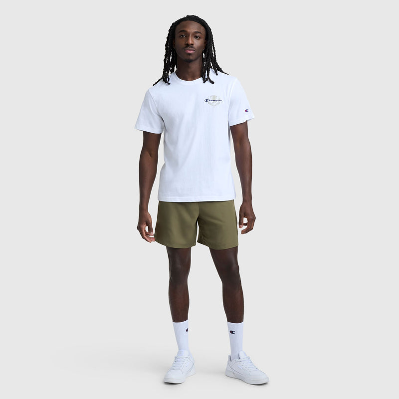 A man with long braided hair stands against a plain light grey background, wearing a Champion Club Graphic T-Shirt, C Logo by Champion, paired with olive green shorts, white socks, and white sneakers.